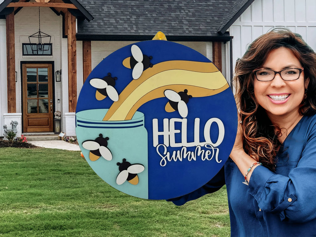 A smiling woman with glasses holds a colorful round sign that says HELLO summer with bees and honey art, standing outside a modern house with a manicured lawn.