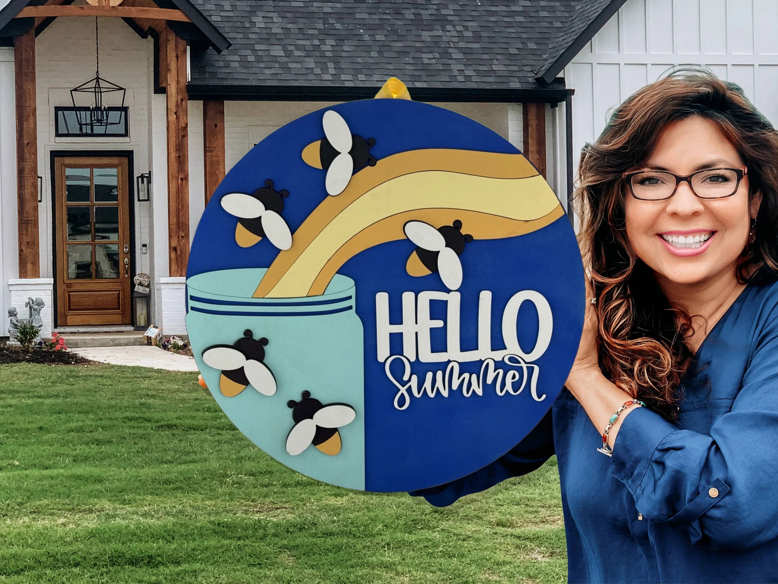 A smiling woman with glasses holds a colorful round sign that says HELLO summer with bees and honey art, standing outside a modern house with a manicured lawn.
