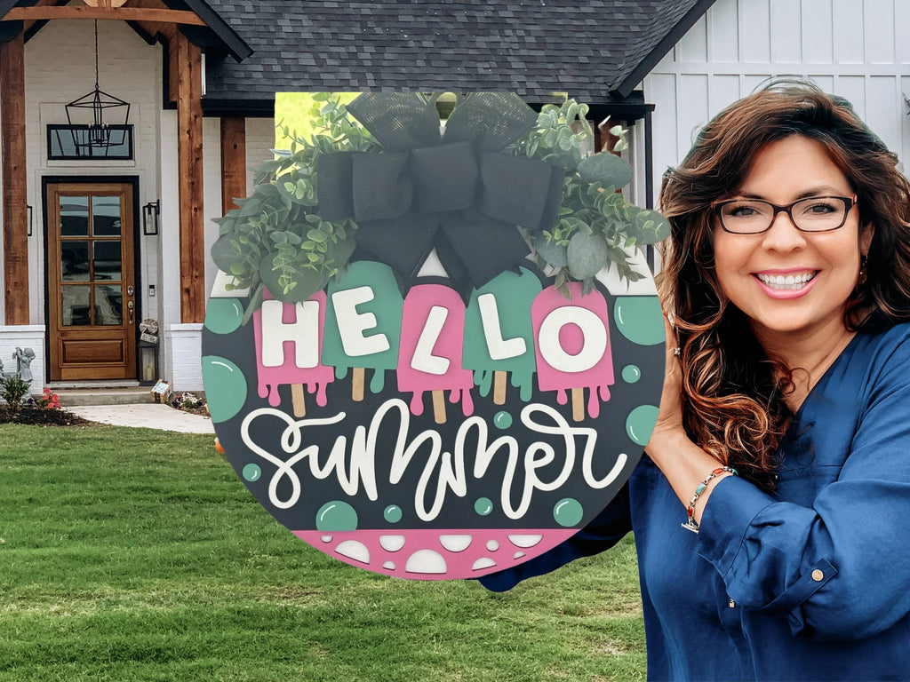 A smiling woman with long brown hair and glasses holds a round sign reading HELLO summer with colorful, playful lettering, greenery, and a black bow. She stands outside a modern house with wood accents, white siding, and a lawn.