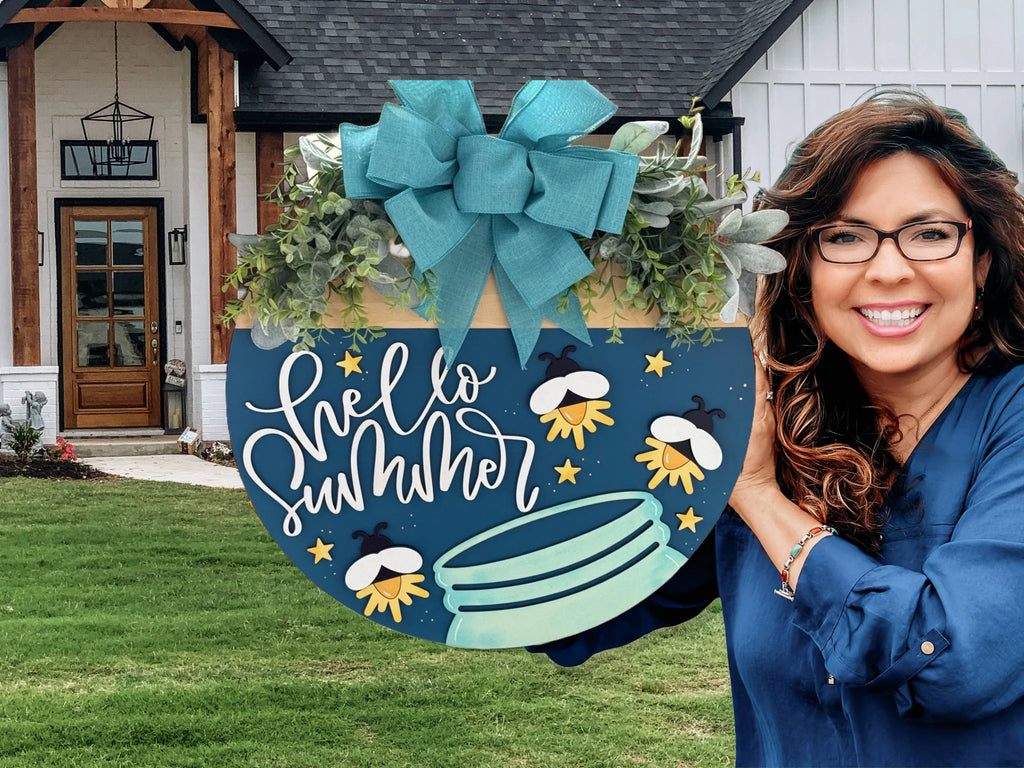 A woman in a blue shirt holds a round sign with Hello Summer written on it. The sign is decorated with a large blue bow, greenery, and illustrations of fireflies, stars, and a jar. A modern house with a lush green lawn is in the background.
