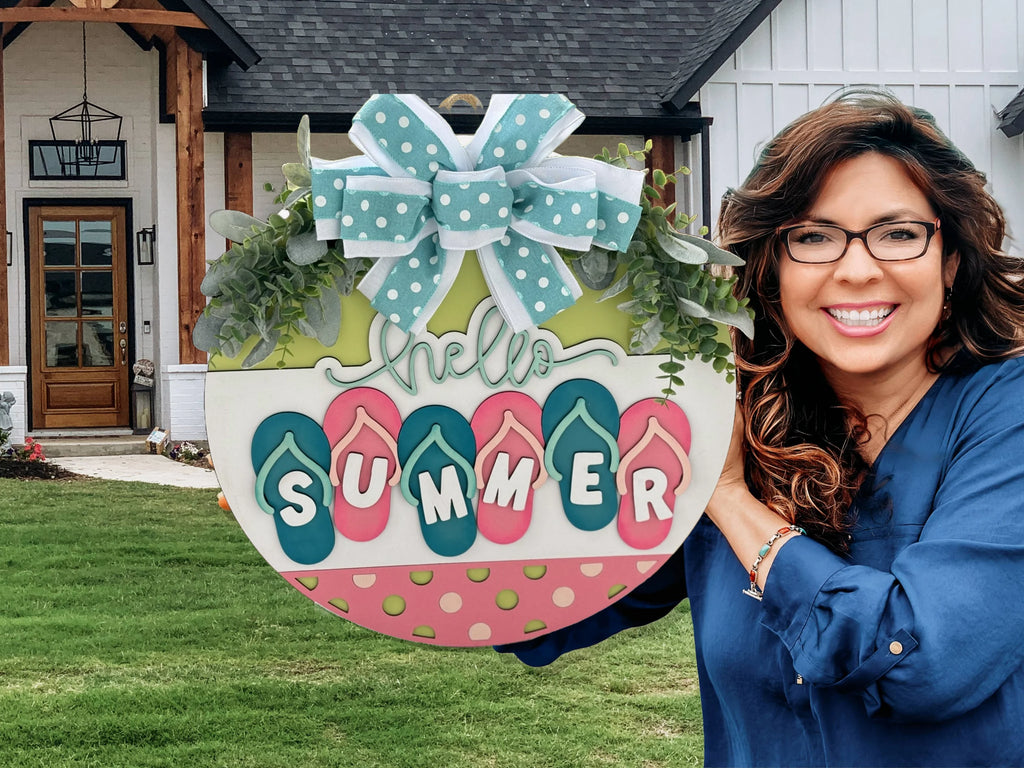 Woman in glasses smiling, holding a colorful round sign that reads Hello Summer with flip-flop decorations. Shes wearing a blue top, standing in front of a house with a large window and wooden beams, next to a well-manicured lawn. The sign is adorned with a polka dot bow.