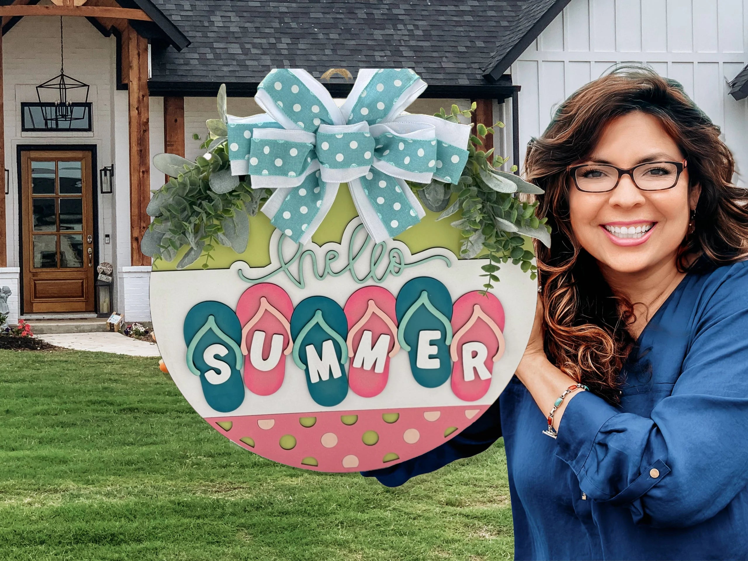 Woman in glasses smiling, holding a colorful round sign that reads Hello Summer with flip-flop decorations. Shes wearing a blue top, standing in front of a house with a large window and wooden beams, next to a well-manicured lawn. The sign is adorned with a polka dot bow.