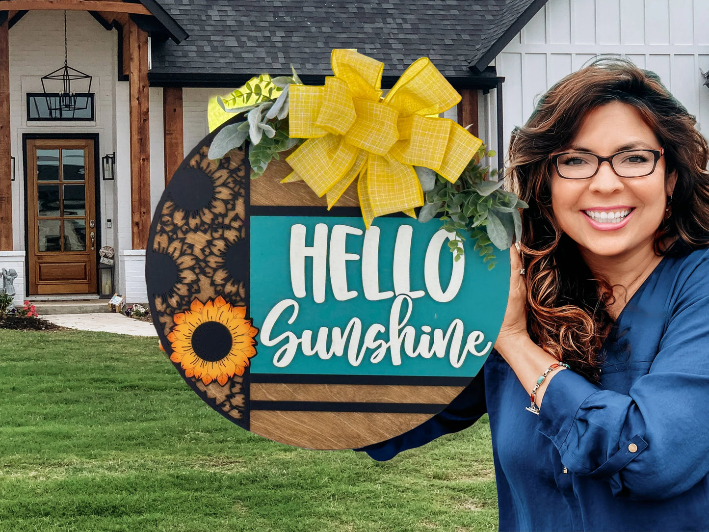 A smiling woman with long brown hair and glasses, wearing a blue shirt, stands outside a modern house holding a large round sign decorated with sunflowers, greenery, a yellow plaid bow, and the words “HELLO Sunshine” in cheerful white and yellow lettering.