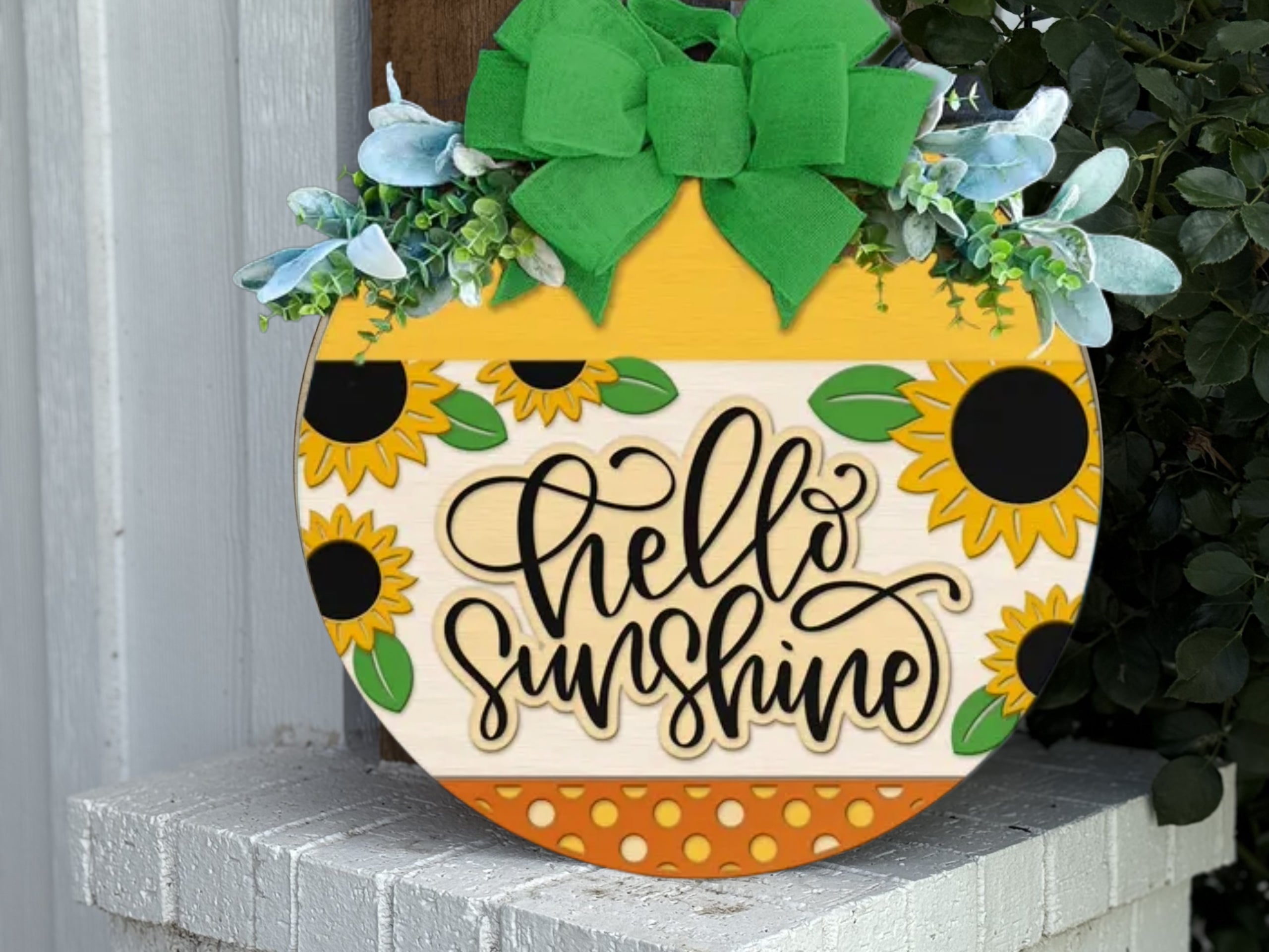Round wooden sign with sunflowers, green leaves, a large green bow, and the words hello sunshine written in script, displayed on a white brick surface near plants.
