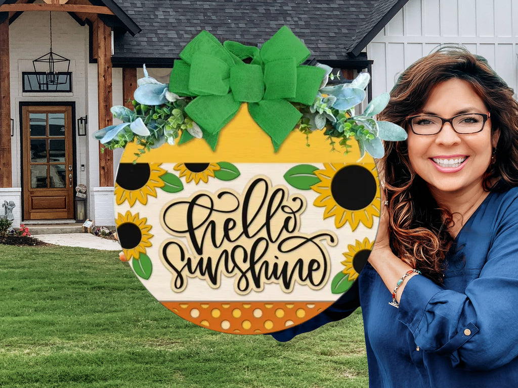 A smiling woman with glasses holds a sunflower-themed sign reading Hello Sunshine in front of a modern house with white siding, wood accents, and a green lawn.