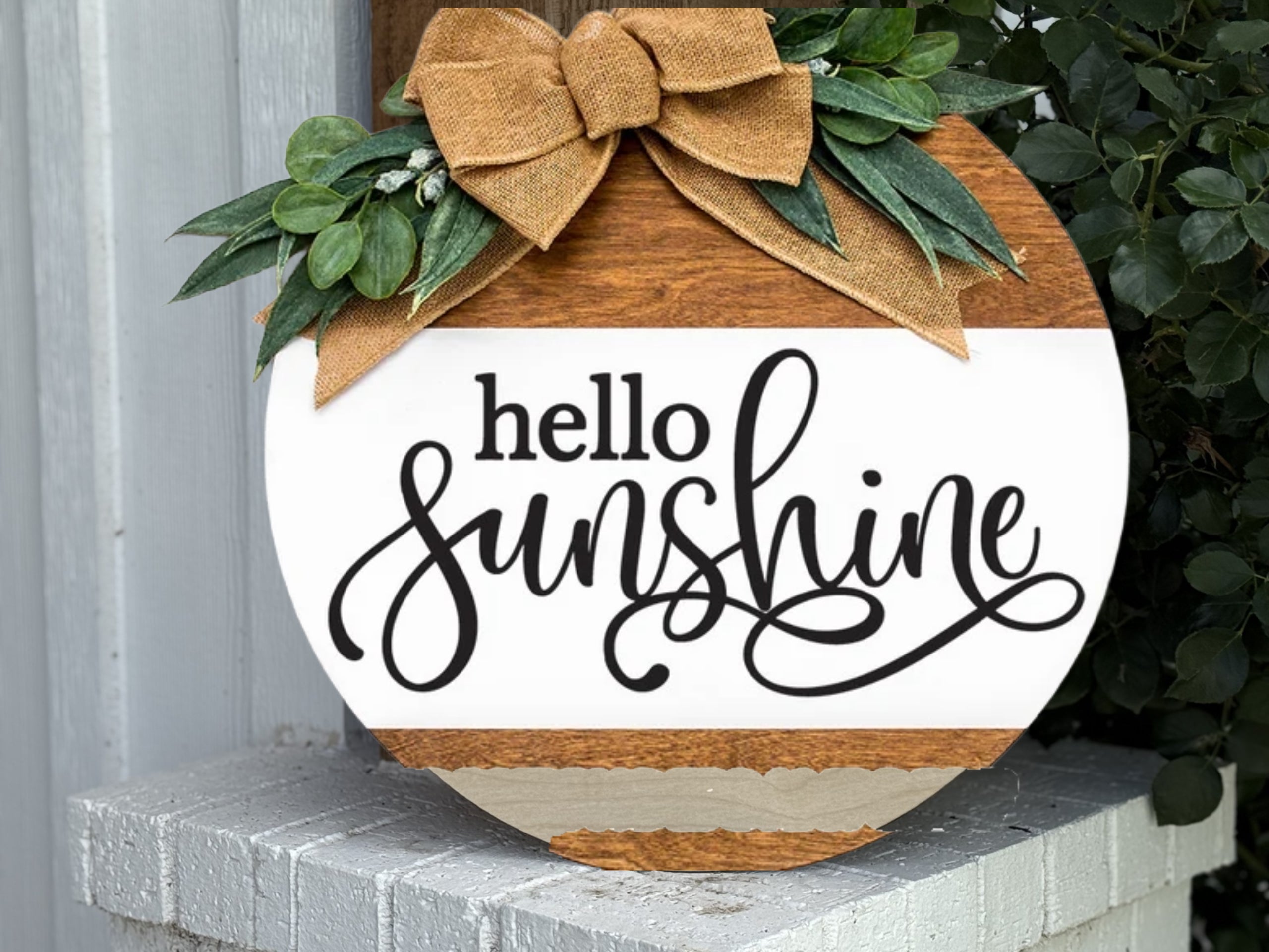 A round wooden sign with greenery and a burlap bow on top, featuring the text hello sunshine in stylish black script. The sign is propped on a white brick surface near some green leaves.
