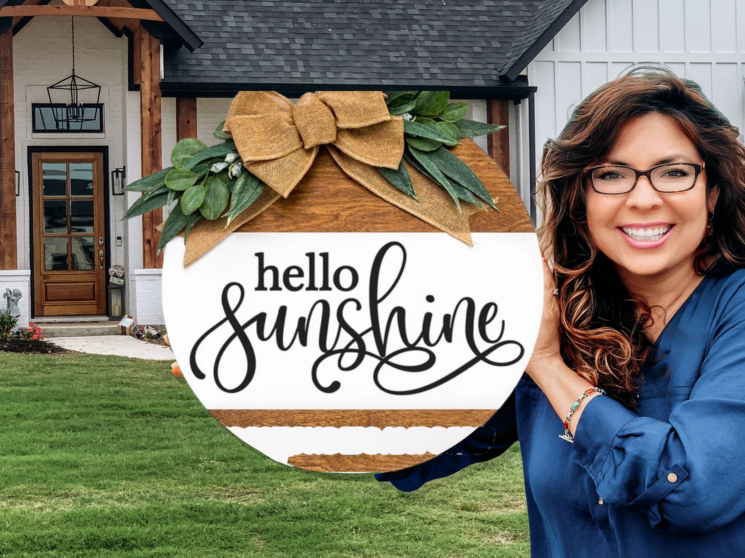 A smiling woman with wavy brown hair and glasses holds a round wooden sign decorated with greenery and a bow that reads hello sunshine in cursive. She stands in front of a modern farmhouse-style home.
