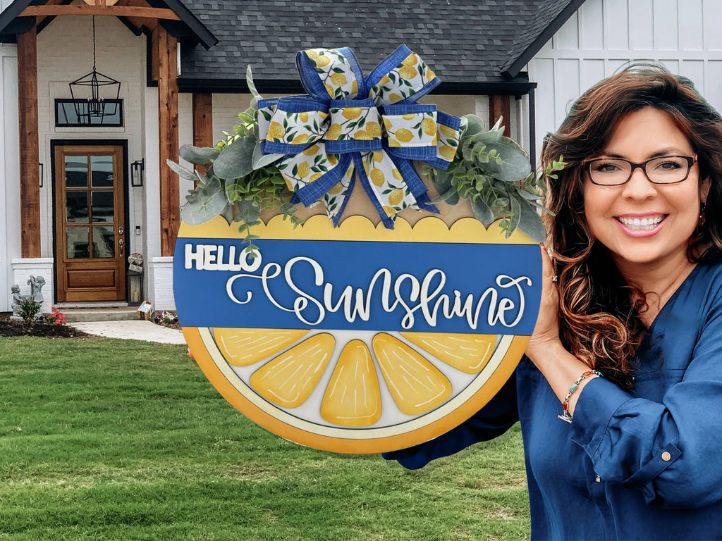 A woman with long brown hair and glasses, wearing a blue blouse, smiles and holds a circular sign that reads Hello Sunshine in white script. The sign features lemon slices, greenery, and a blue and yellow bow. A white house with brown trim and a front porch is in the background.
