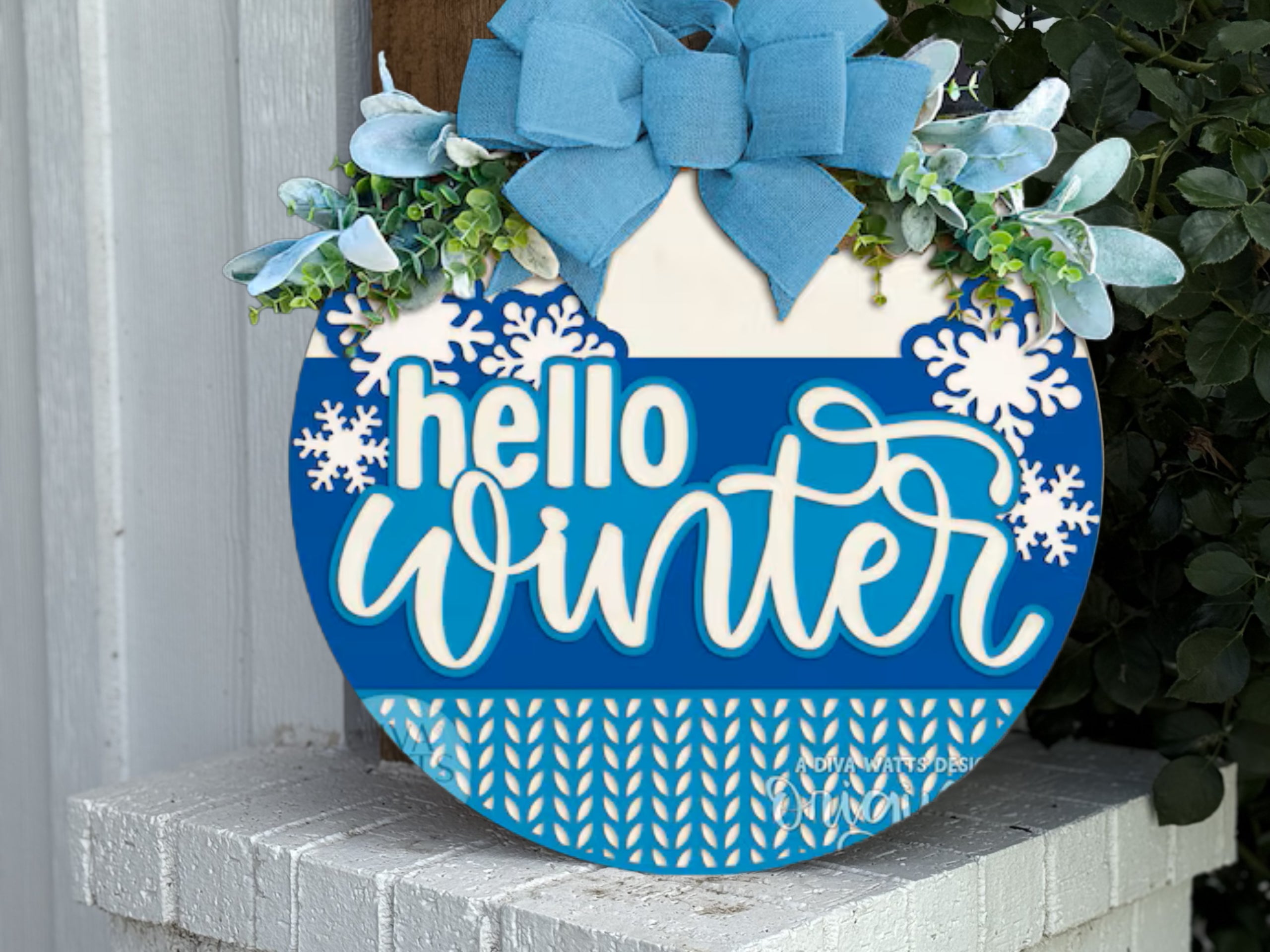 A round blue and white sign with a large blue bow and greenery on top reads hello winter with snowflake decorations, leaning against a white brick wall.