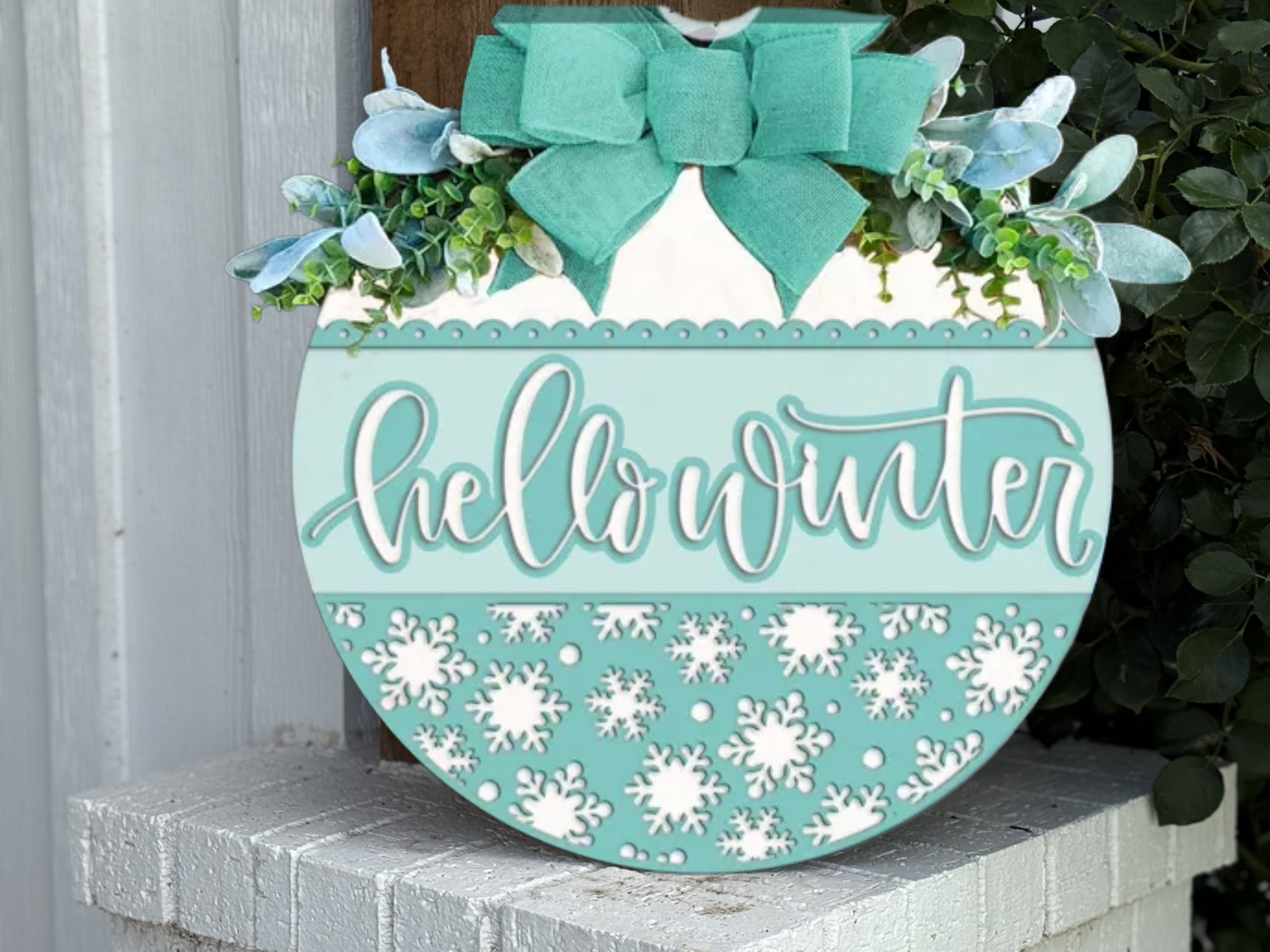 A round teal and white sign with a green bow, greenery, and the words hello winter in script, decorated with white snowflakes, hanging on a white brick wall.