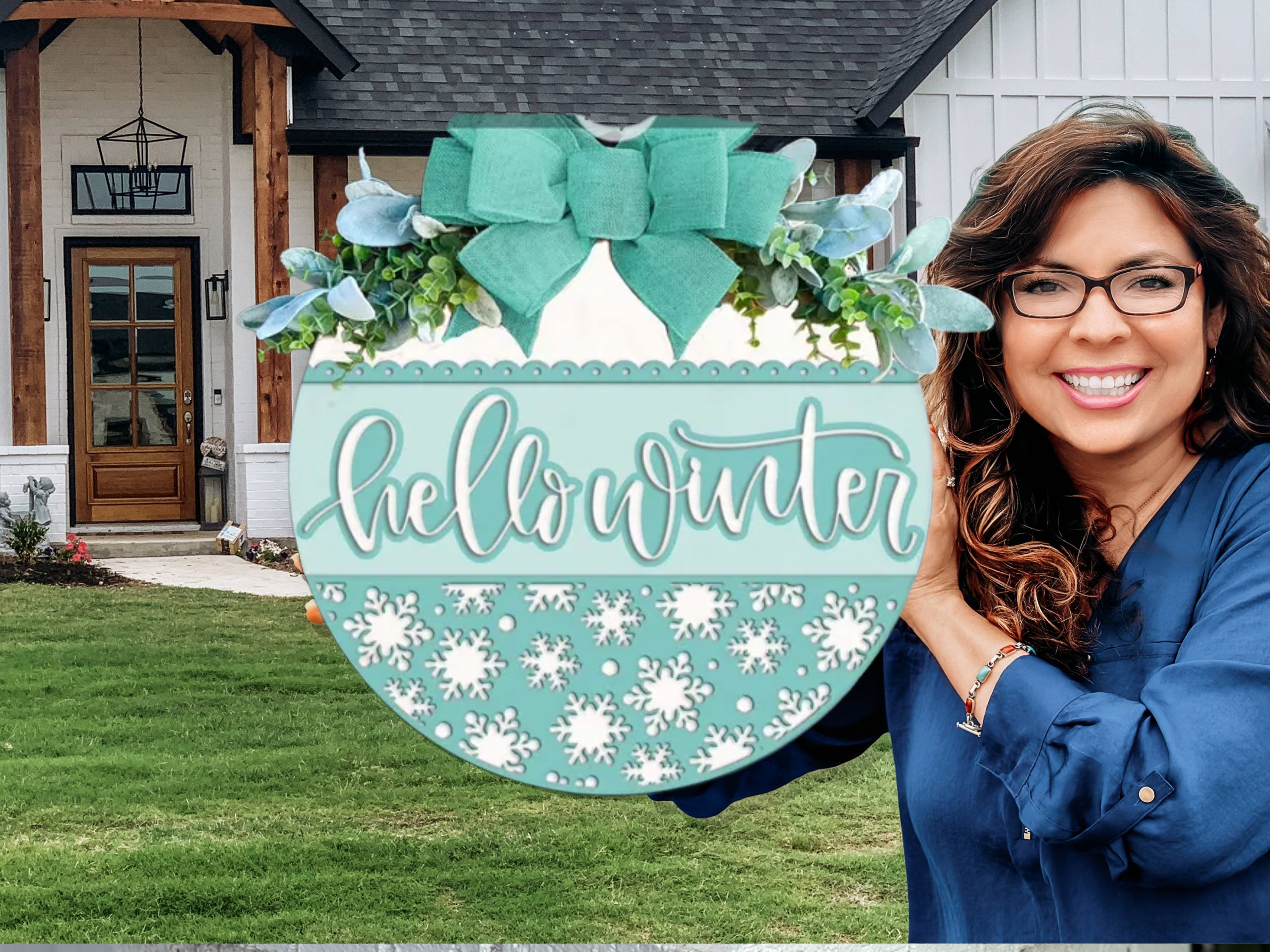 A smiling woman with long brown hair and glasses holds a large round teal sign decorated with leaves and a bow. The sign says hello winter with snowflake designs. She stands in front of a modern house with a lawn.