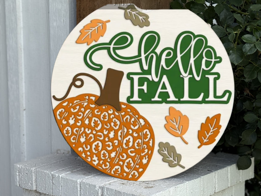 A woman with long brown hair and glasses smiles while holding a round sign that says hello FALL with a decorative pumpkin and autumn leaves, standing in front of a modern house.