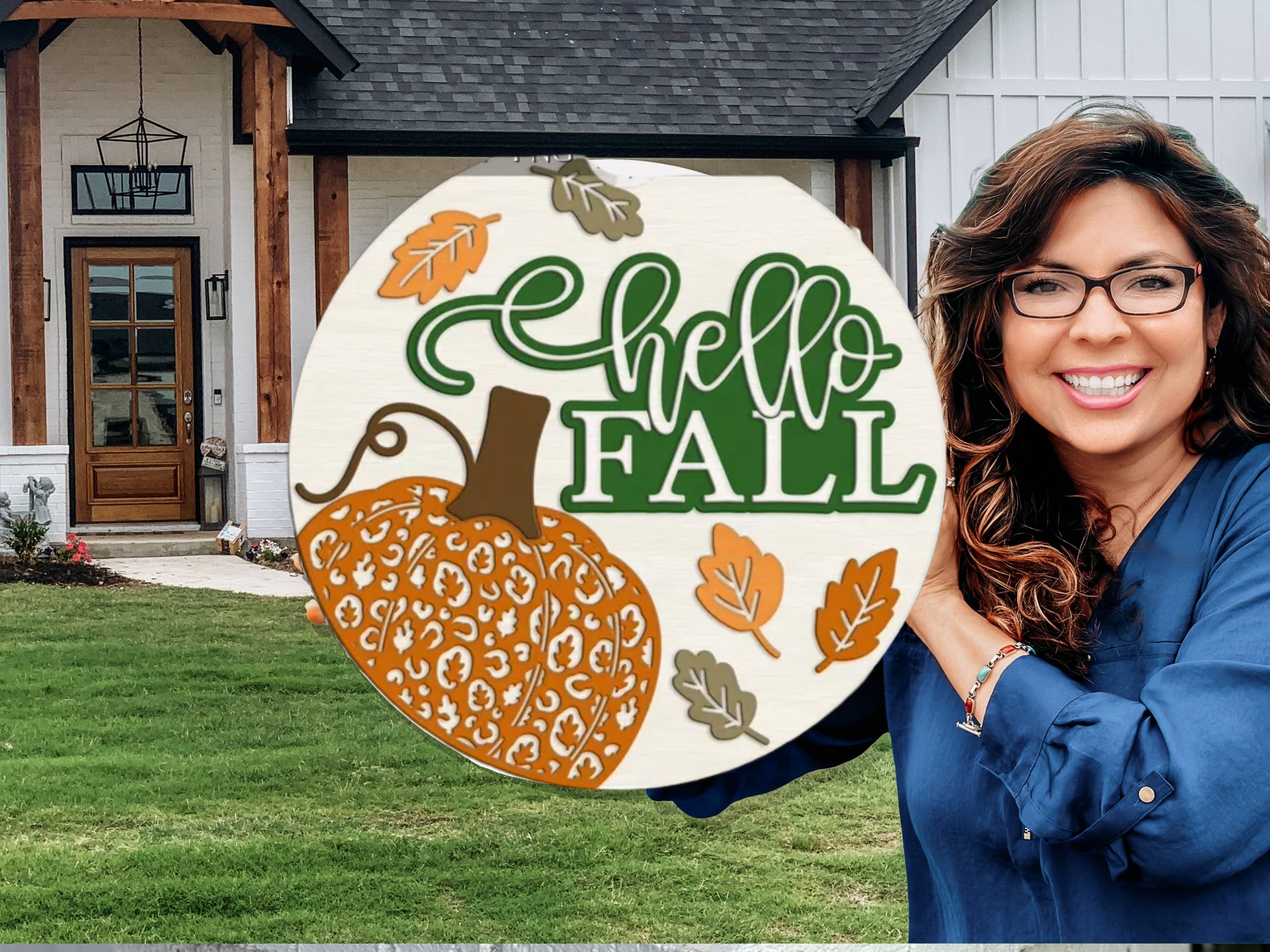 A woman with long brown hair and glasses smiles while holding a round sign that says hello FALL with a decorative pumpkin and autumn leaves, standing in front of a modern house.