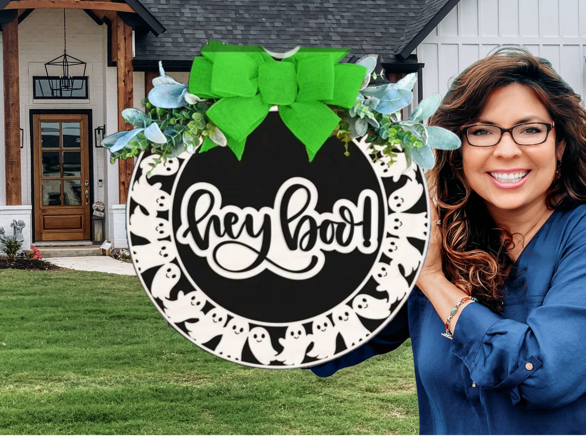 A smiling woman with long brown hair and glasses holds a round sign with a green bow and “hey boo!” written in white script, standing in front of a modern house with a green lawn. The sign is decorated with ghost illustrations.