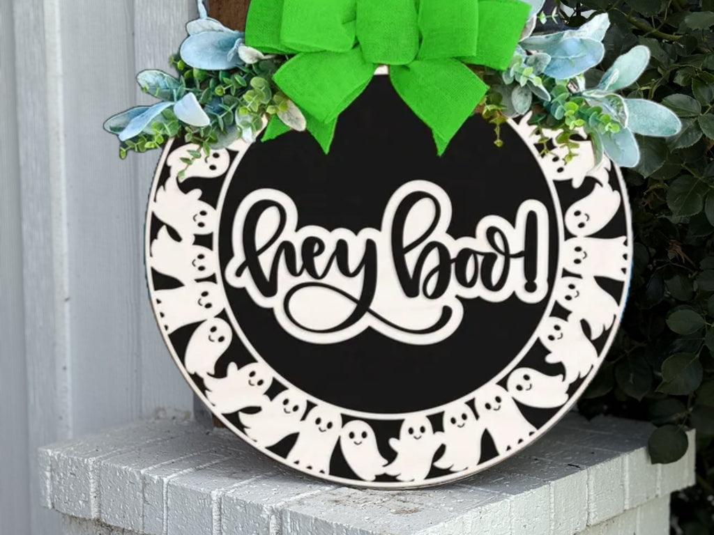 A round sign decorated with a green bow and greenery reads hey boo! in cursive. The border features a pattern of white cartoon ghosts on a black background. The sign is propped on a white brick surface.