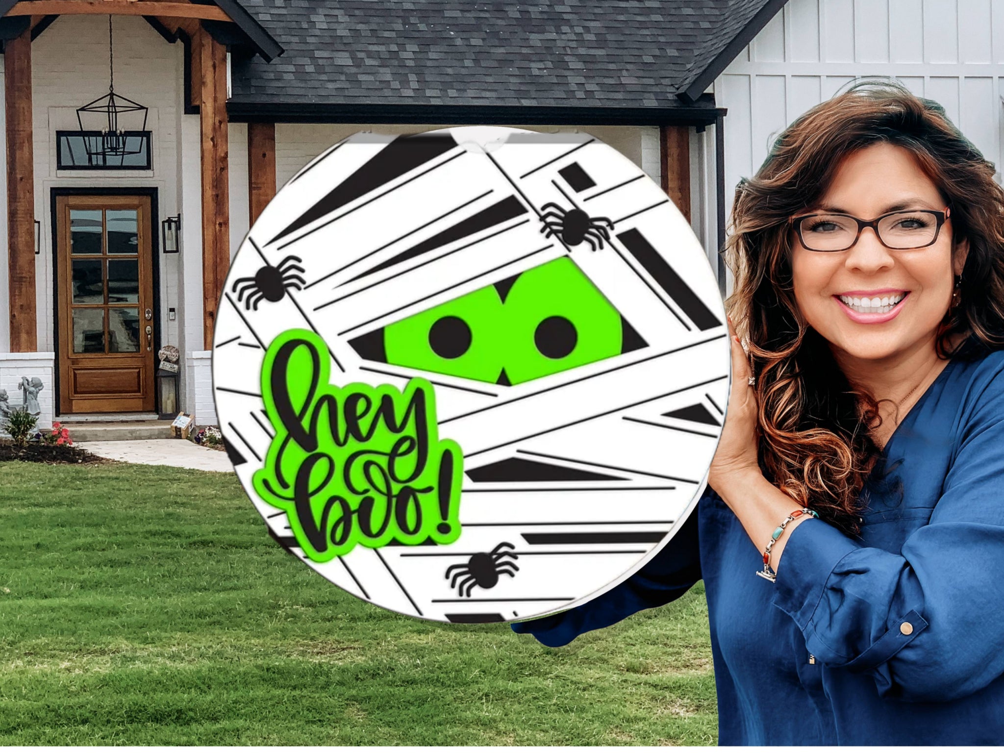A woman with long brown hair and glasses holds a round Halloween sign featuring green eyes peeking through mummy bandages, small black spiders, and the words “hey boo!” in green. A modern house is in the background.