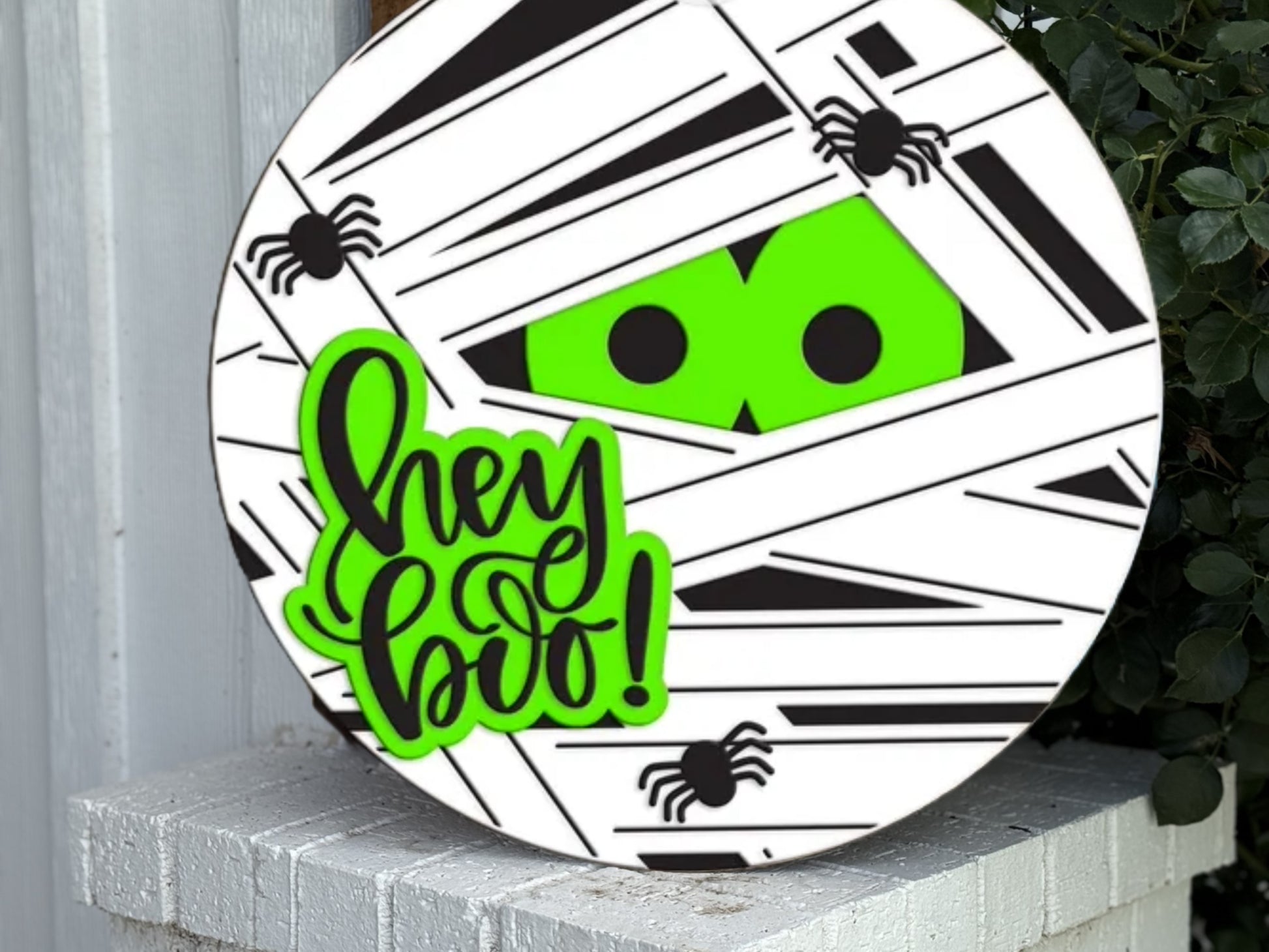 A round Halloween sign with a mummy design, bright green eyes peeking through white and black wrappings, small black spiders, and the phrase “hey boo!” in green and black lettering.