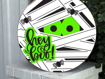 A round Halloween sign with a mummy design, bright green eyes peeking through white and black wrappings, small black spiders, and the phrase “hey boo!” in green and black lettering.