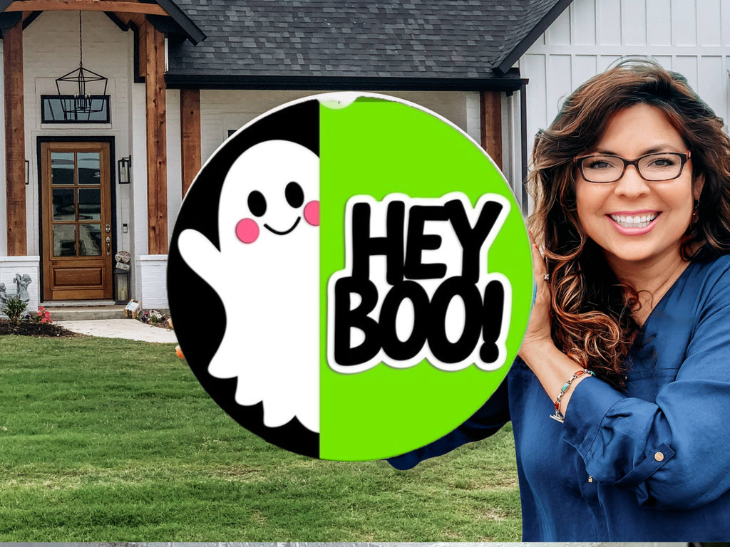A smiling woman holding a round sign with a cute ghost and the words HEY BOO! in front of a modern house with a green lawn.