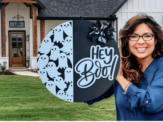 A round black and white Halloween sign features cartoon ghosts on one side, a Hey Boo! message in bold script on the other, and a bow with ghost patterns at the top. The sign is propped against a white brick wall.