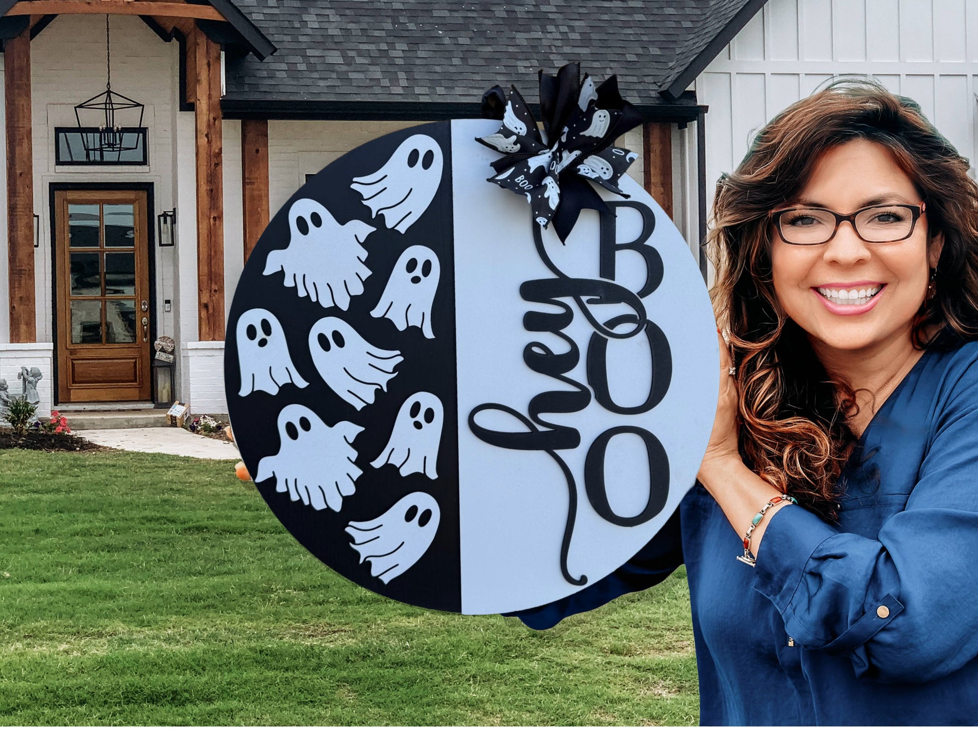 A smiling woman with long brown hair and glasses holds a round sign decorated with cartoon ghosts, the word “hey,” and large letters spelling “BOO,” standing in front of a modern house with a green lawn.
