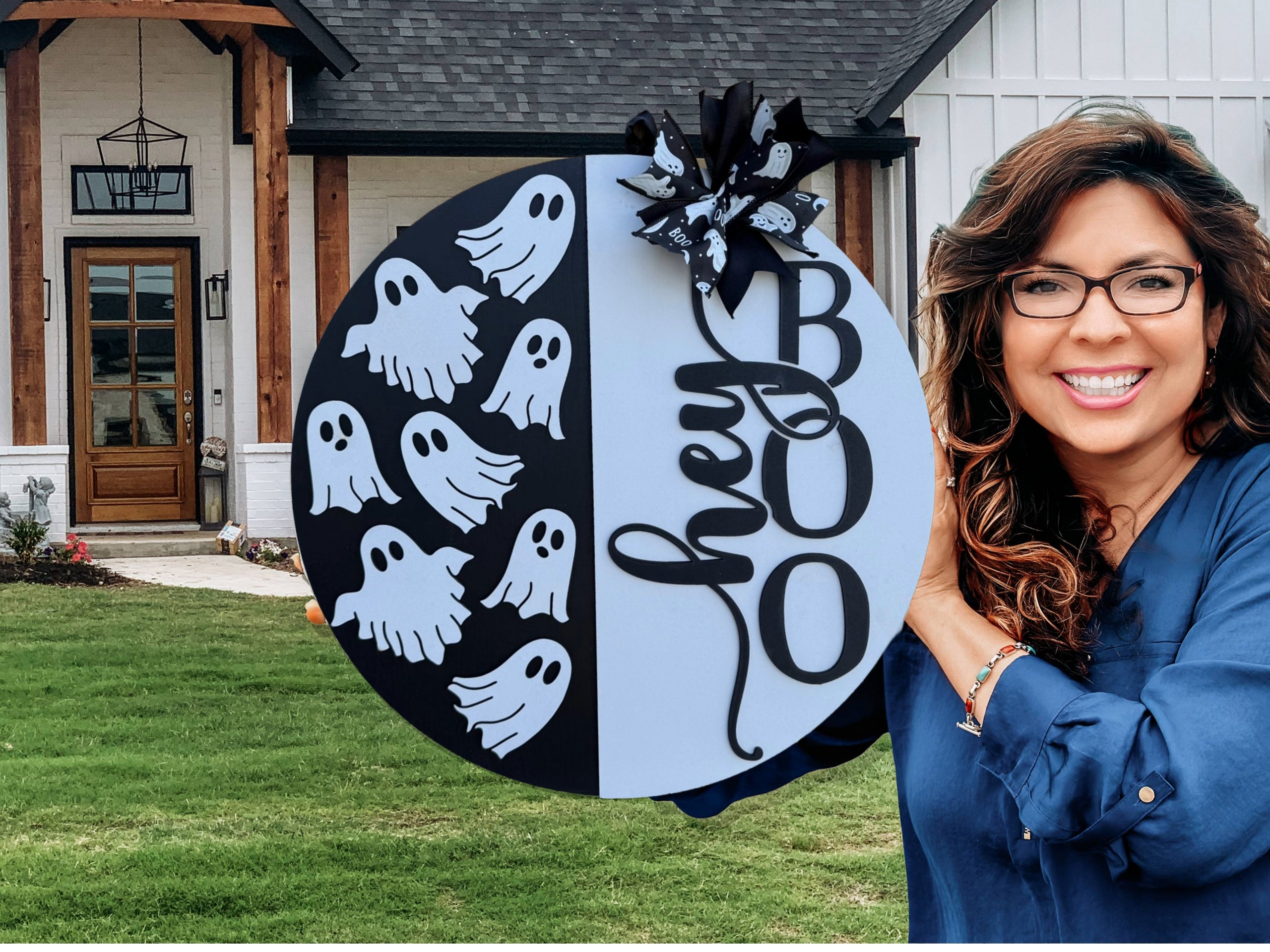 A smiling woman with long brown hair and glasses holds a round sign decorated with cartoon ghosts, the word “hey,” and large letters spelling “BOO,” standing in front of a modern house with a green lawn.