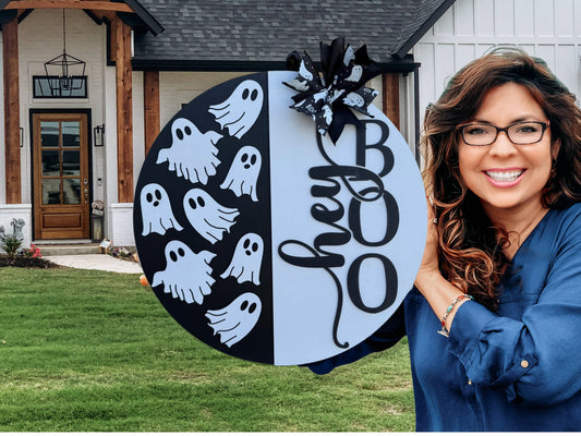 A smiling woman with long brown hair and glasses holds a round sign decorated with cartoon ghosts, the word “hey,” and large letters spelling “BOO,” standing in front of a modern house with a green lawn.