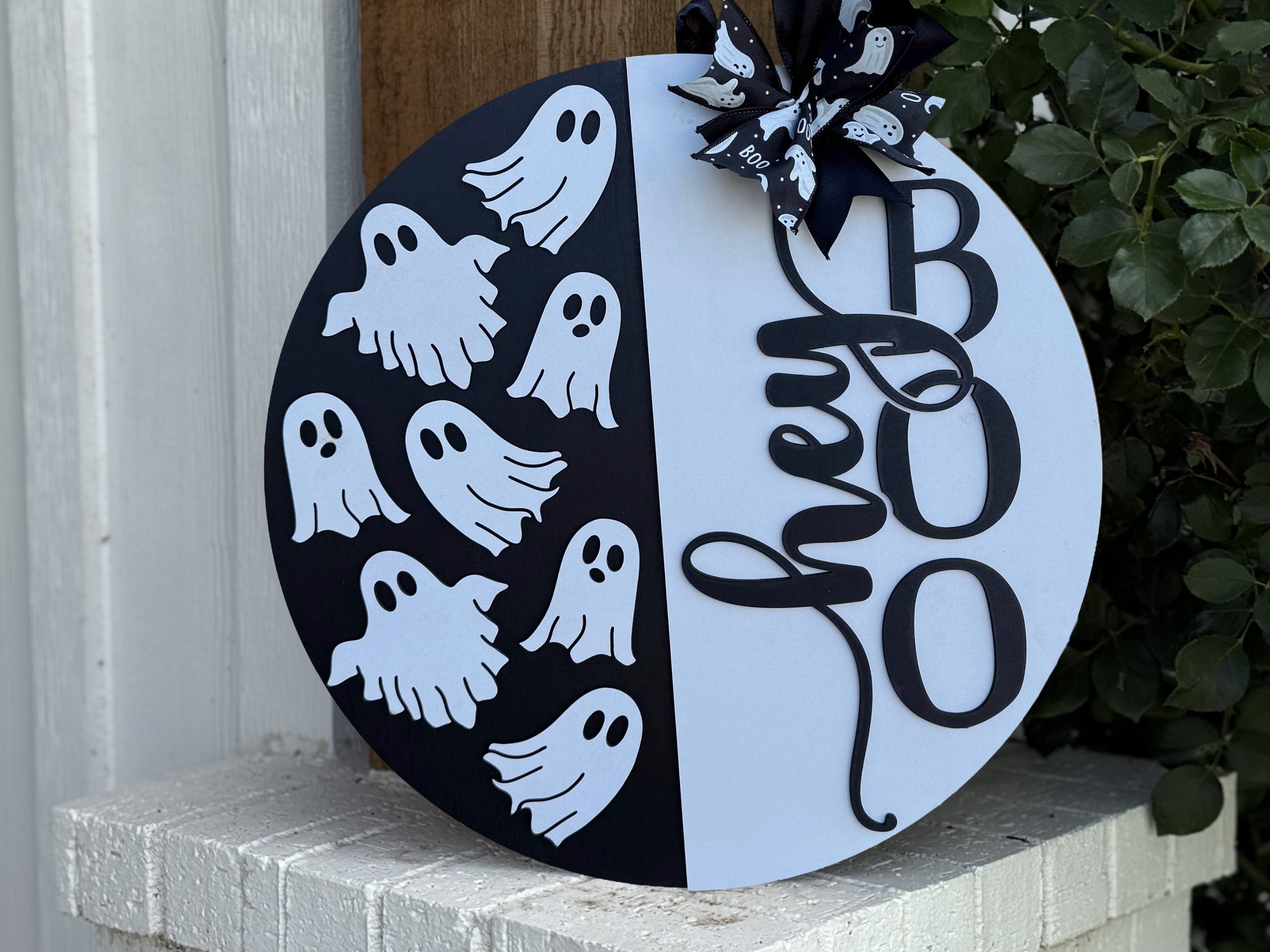 A round Halloween sign split into black and white halves, featuring cutout ghosts on the left and the words hey BOO in bold black letters on the right, topped with a black ribbon with white ghosts.
