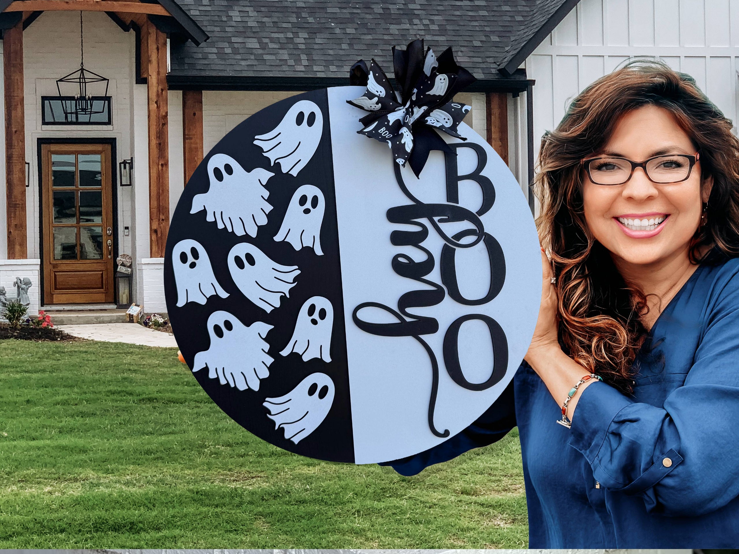 A smiling woman stands outside a house holding a large round Halloween sign decorated with cartoon ghosts and the words hey BOO in black lettering, topped with a black and white bow.