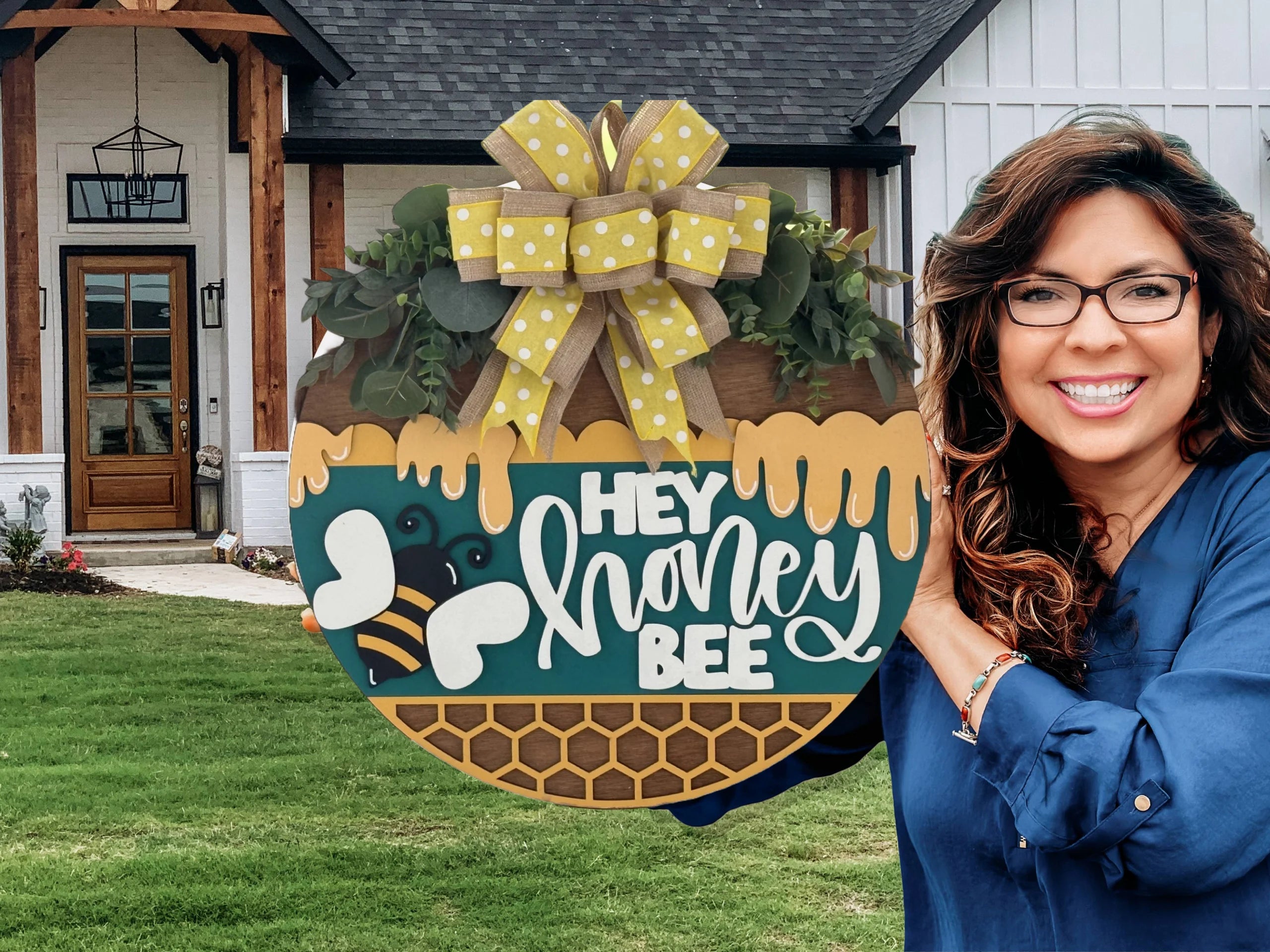 A smiling woman with wavy brown hair and glasses holds a round wooden sign decorated with yellow ribbon, greenery, bees, and honeycomb patterns. The sign says “HEY honey BEE.” A modern house with a lawn and wooden door is in the background.