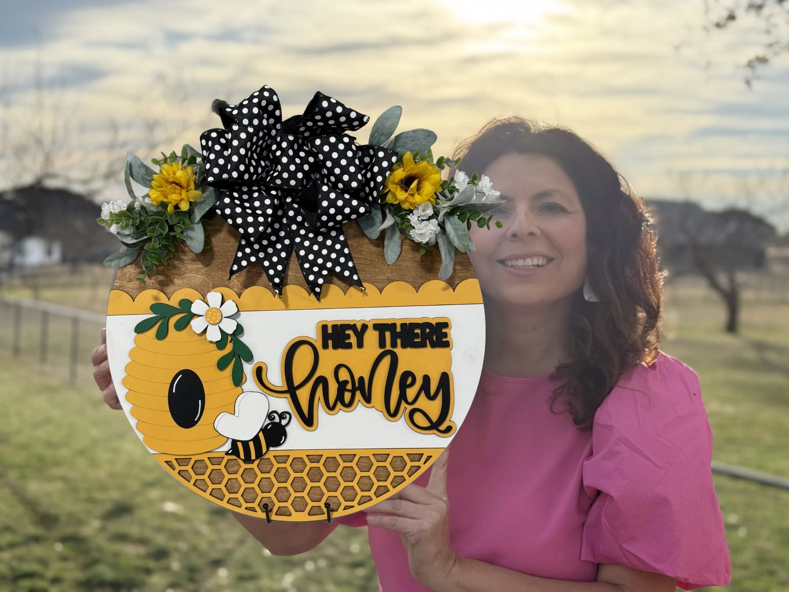 A woman in a pink blouse holds a round, bee-themed decorative sign. The sign features a beehive, flowers, and a bee with the text Hey There Honey in playful yellow and black script. It is adorned with a black and white polka dot bow and sunflowers. The sky is partly cloudy.