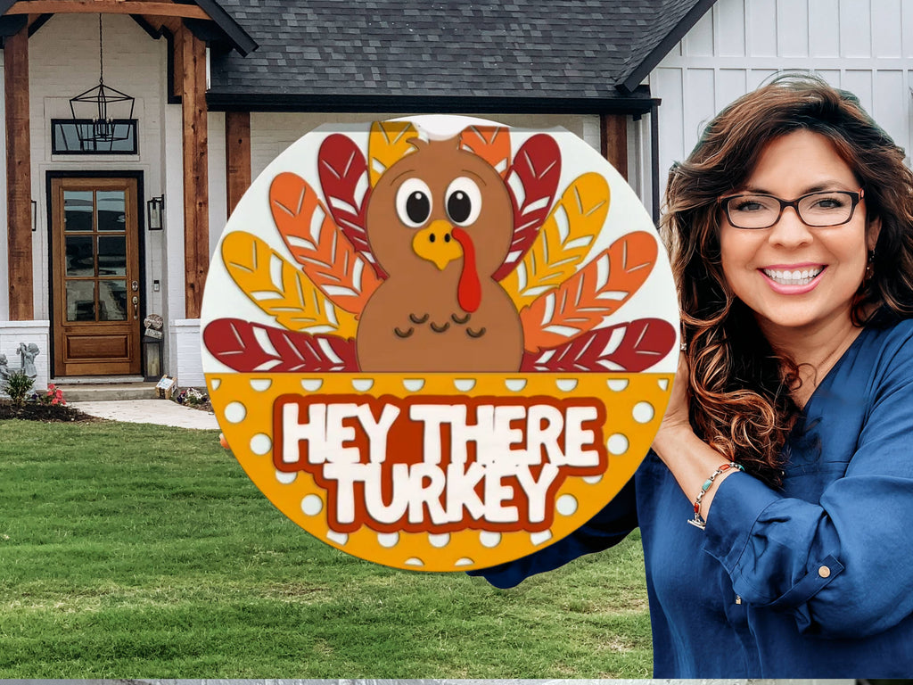A smiling woman stands outside a house, holding a colorful round sign with a cartoon turkey and the words HEY THERE TURKEY in bold white letters. The house and green lawn are visible in the background.