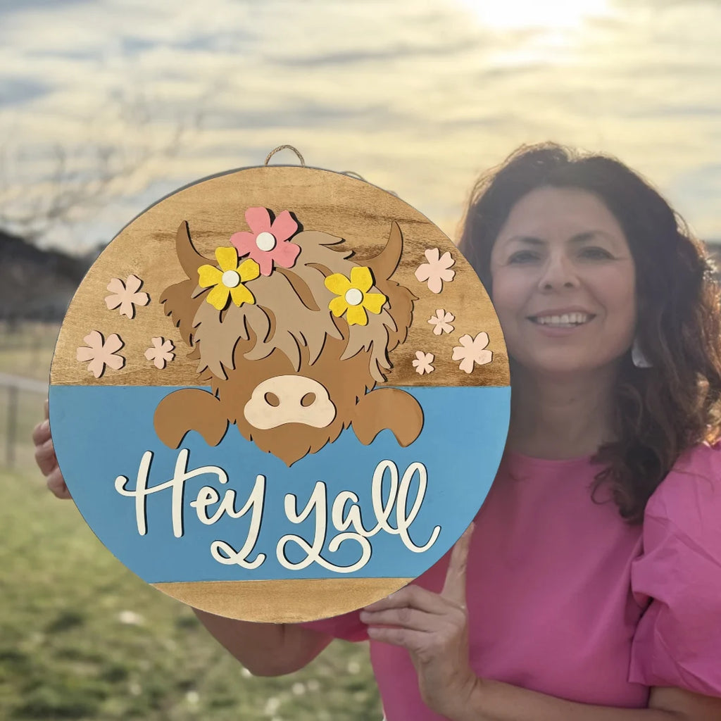 Woman in a pink top holds a round wooden sign with a cartoon cow wearing flowers. The sign has a blue bottom half with Hey yall written in white script. A grassy field and cloudy sky are in the background.