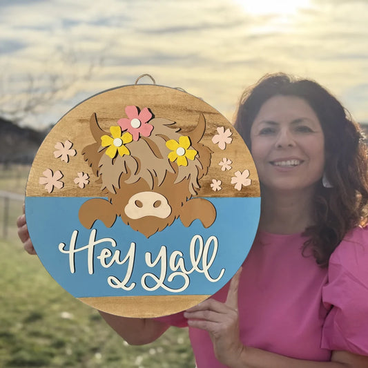 Woman in a pink top holds a round wooden sign with a cartoon cow wearing flowers. The sign has a blue bottom half with Hey yall written in white script. A grassy field and cloudy sky are in the background.