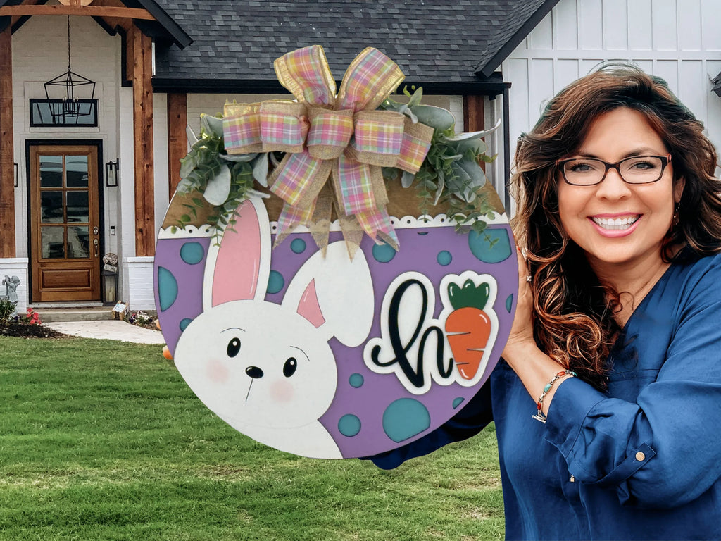 A smiling woman with long brown hair and glasses holds a round Easter-themed sign with a bunny, polka dots, greenery, a bow, and the word hi with a carrot. She stands outside in front of a modern house with a lawn and wooden beams by the door.