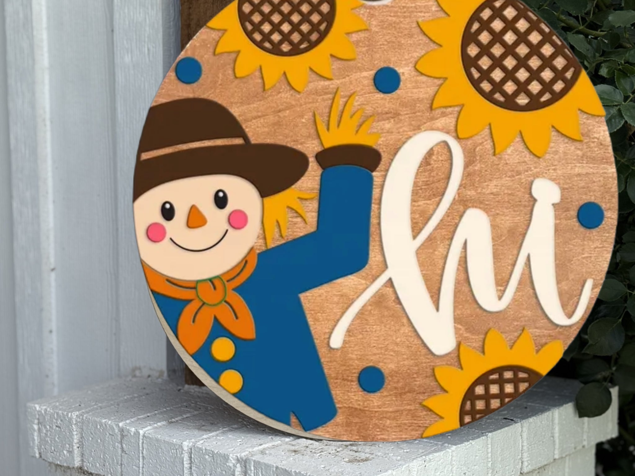 A round wooden sign with a cartoon scarecrow waving, surrounded by sunflowers and blue dots, and the word hi in large white script. The sign is placed on a white brick ledge by green foliage.