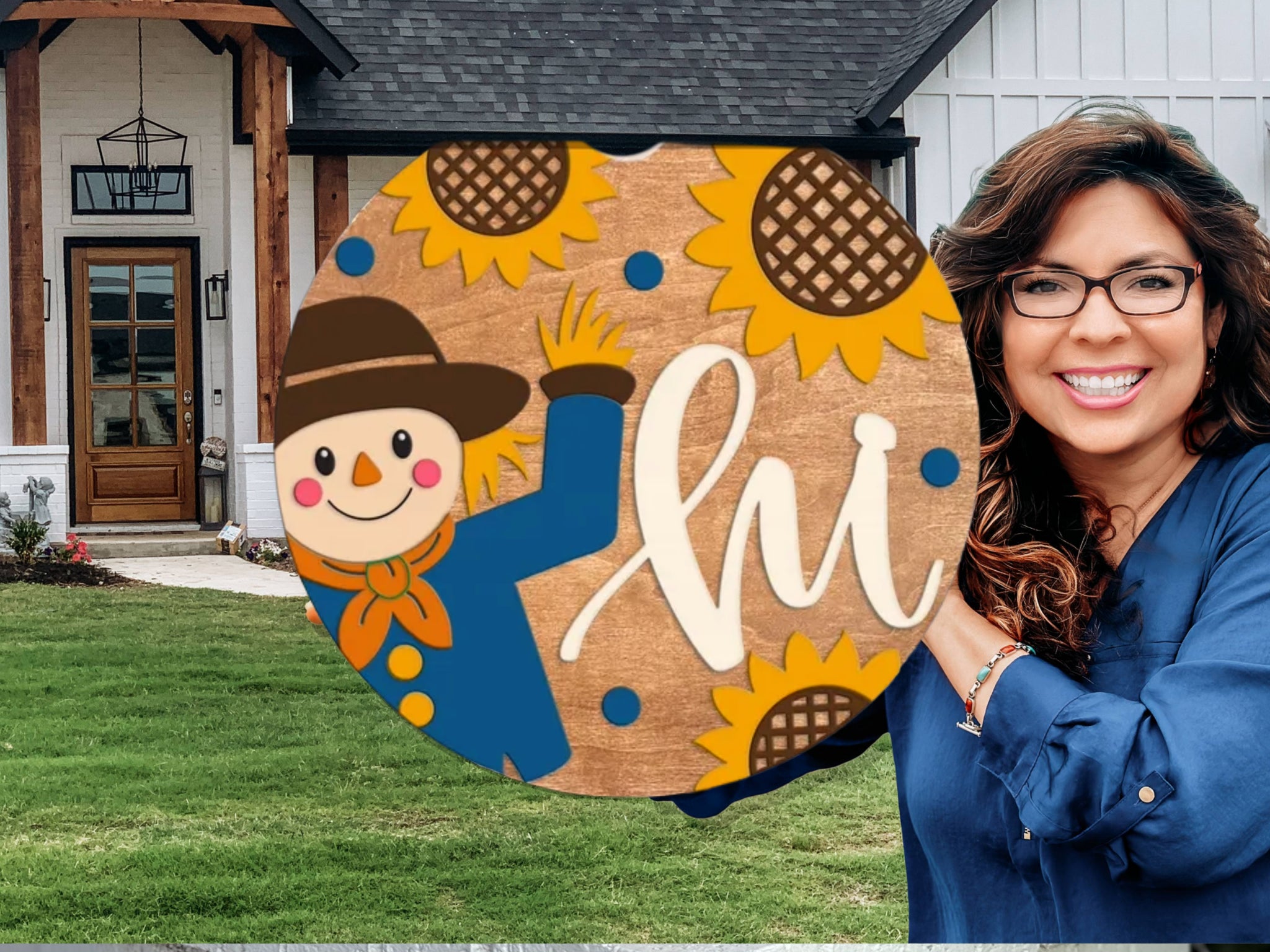 A smiling woman holding a round wooden sign with a cartoon scarecrow, yellow sunflowers, and the word hi in white script, standing in front of a modern house with a dark roof and white siding.