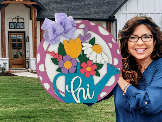 A smiling woman with wavy brown hair and glasses holds a round, floral welcome sign that says “hi” in front of a modern house with wood and white accents and a neatly manicured lawn. The sign features colorful flowers and a purple bow.