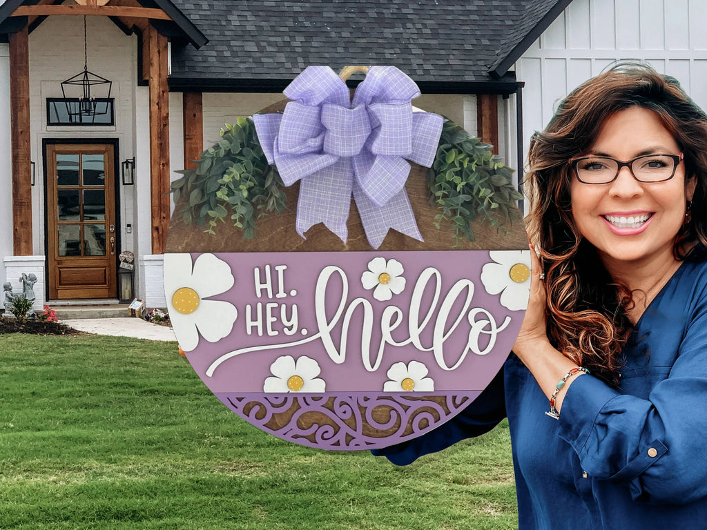 A smiling woman with wavy brown hair and glasses holds a round, purple welcome sign that says “Hi. Hey. Hello” with daisies and a large purple bow. She stands in front of a modern farmhouse with white siding, black trim, and a wooden porch.
