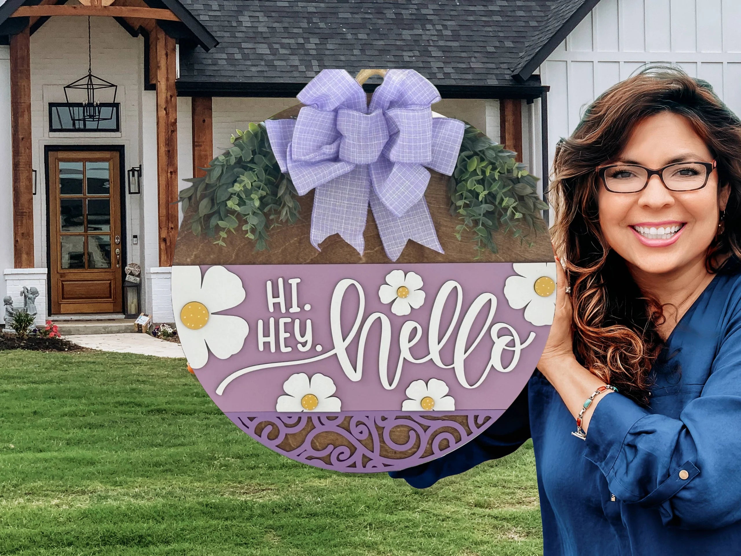 A smiling woman with wavy brown hair and glasses holds a round, purple welcome sign that says “Hi. Hey. Hello” with daisies and a large purple bow. She stands in front of a modern farmhouse with white siding, black trim, and a wooden porch.