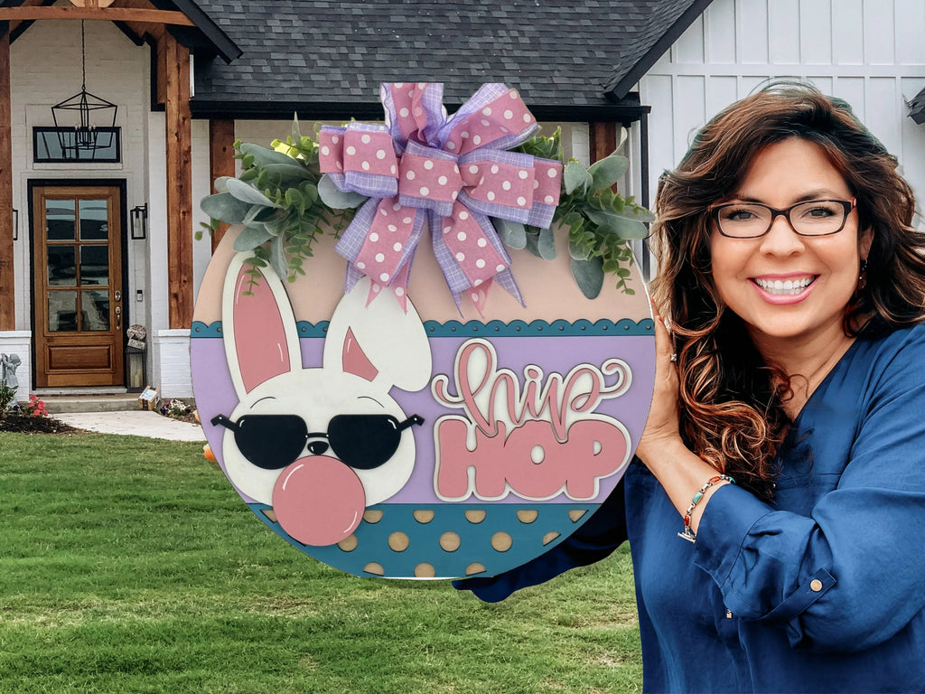 A smiling woman with long, wavy brown hair and glasses holds a round sign decorated with a bunny wearing sunglasses, a pink bow, and the words “hip hop.” The sign is pastel-colored with polka dots and topped with a purple bow. A modern house is in the background.