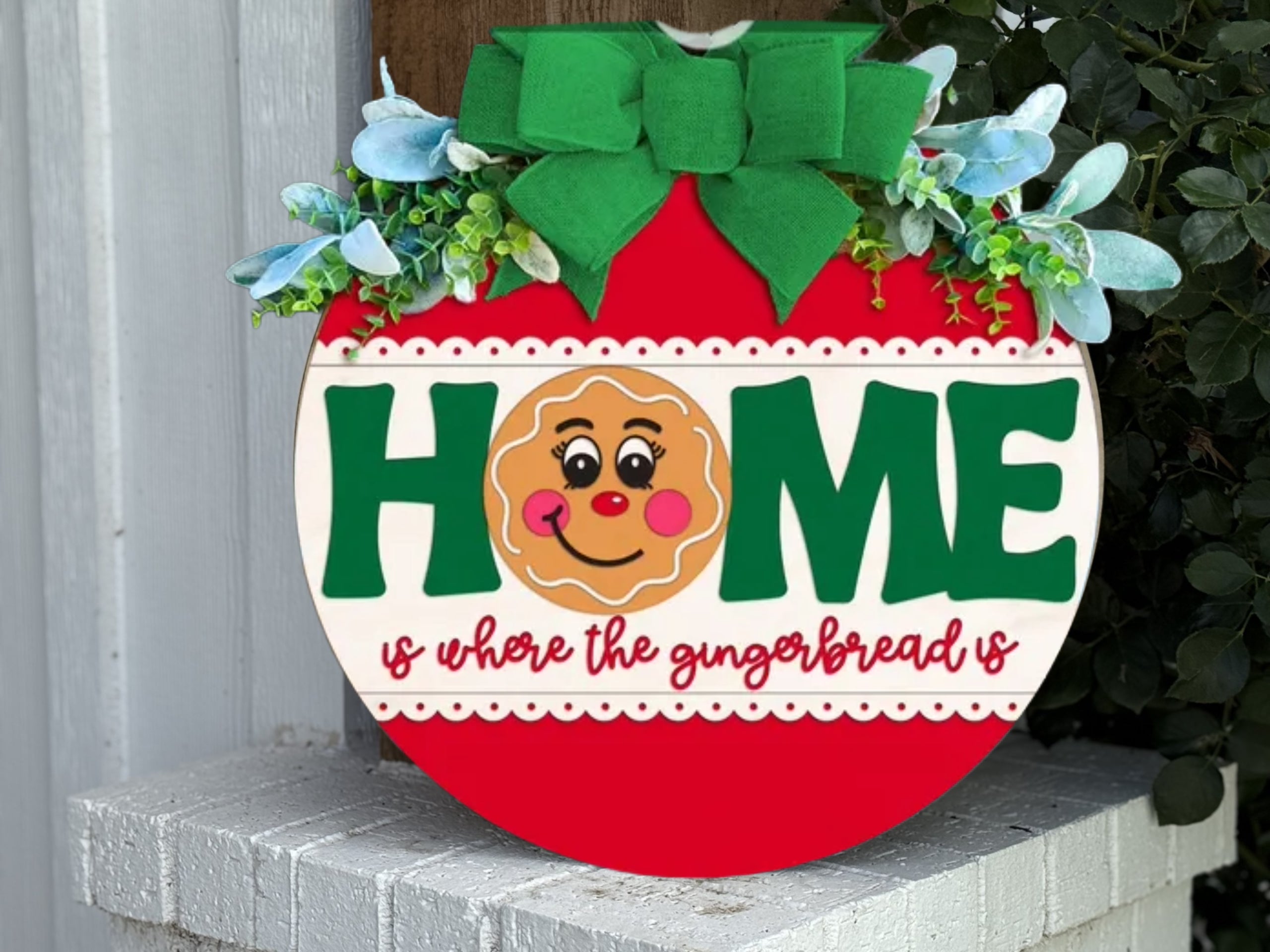 A round sign decorated with greenery and a green bow reads HOME with a smiling gingerbread face as the O. Below, it says, is where the gingerbread is in red script. The sign rests on a white brick ledge.