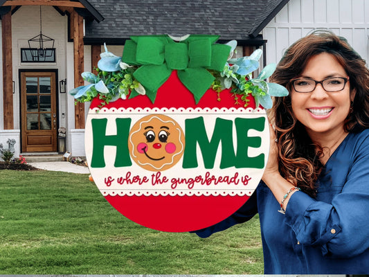 A round sign decorated with greenery and a green bow reads HOME with a smiling gingerbread face as the O. Below, it says, is where the gingerbread is in red script. The sign rests on a white brick ledge.