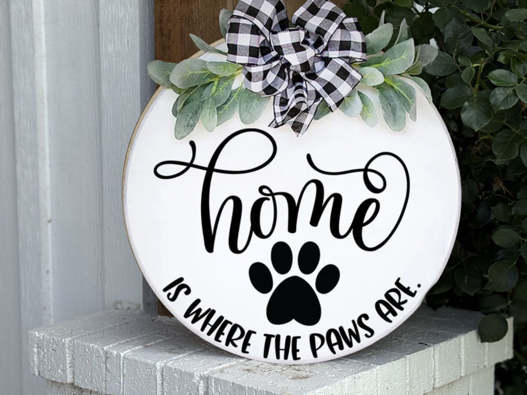 A round white sign with a black paw print, greenery, and a black-and-white plaid bow reads Home is where the paws are. The sign is displayed on a white brick surface.