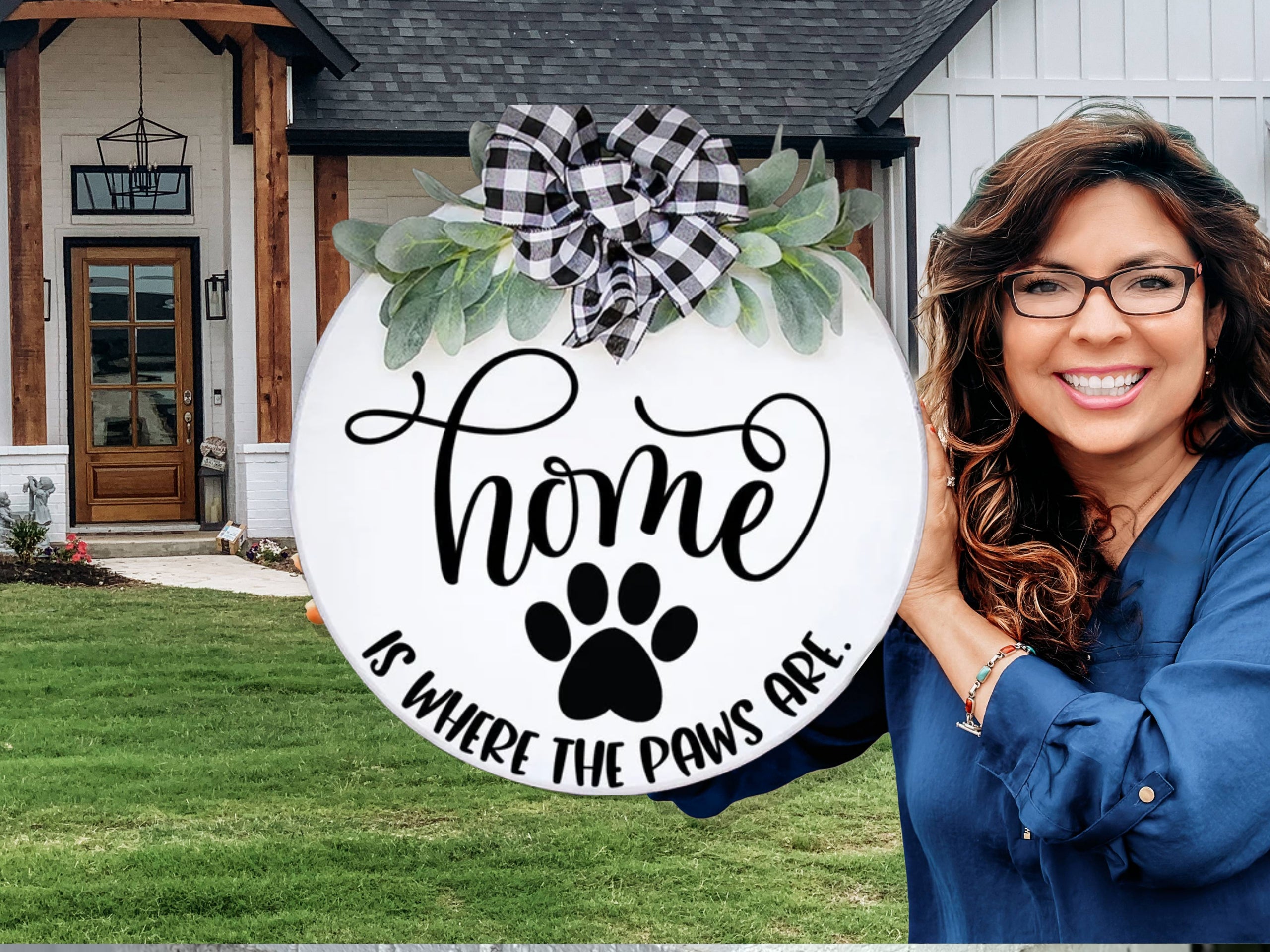 A smiling woman holds a round sign decorated with greenery and a plaid bow, reading Home is where the paws are, featuring a paw print. She stands outside a modern house with a lawn and wooden accents.