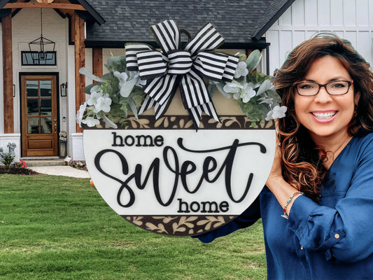 A smiling woman with wavy hair and glasses holds a round “Home Sweet Home” sign with black-and-white stripes, florals, and a bow. She stands outside a modern house with white siding, wood accents, and a green lawn.