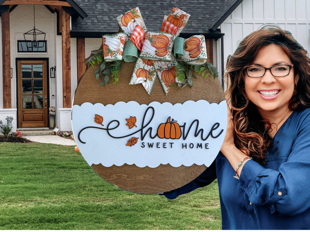 A round wooden sign with a white scalloped section reads Home Sweet Home, with a pumpkin replacing the o in home. It’s decorated with a fall-themed bow and greenery at the top.