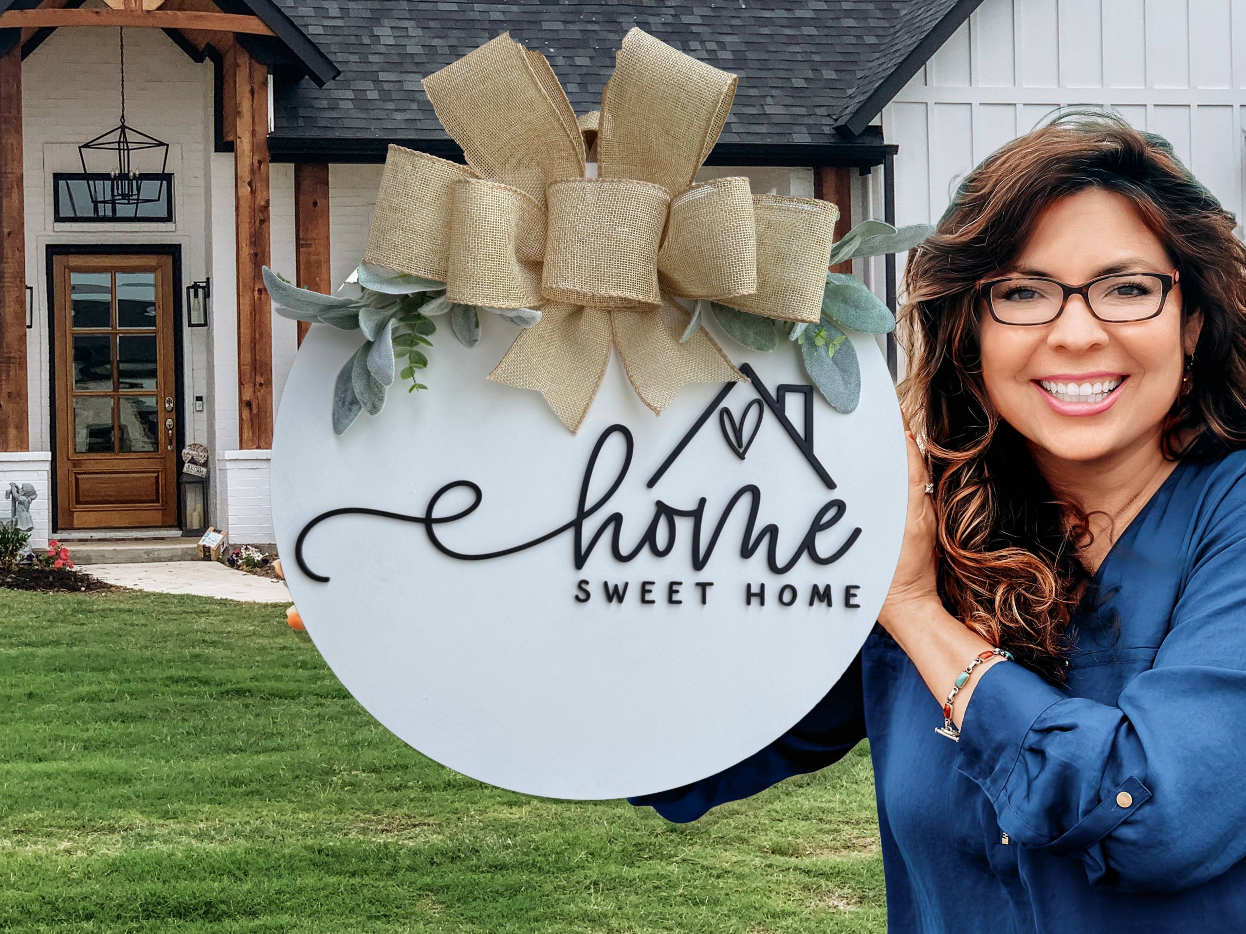 A smiling woman with wavy brown hair and glasses holds a round Home Sweet Home sign with a decorative bow and greenery, standing in front of a modern farmhouse-style home with a wooden porch.