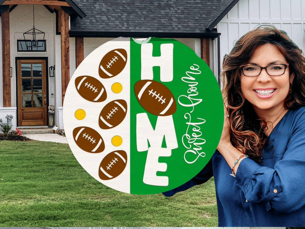 A smiling woman stands in front of a house holding a round sign decorated with footballs and the words HOME Sweet Home in white and green.