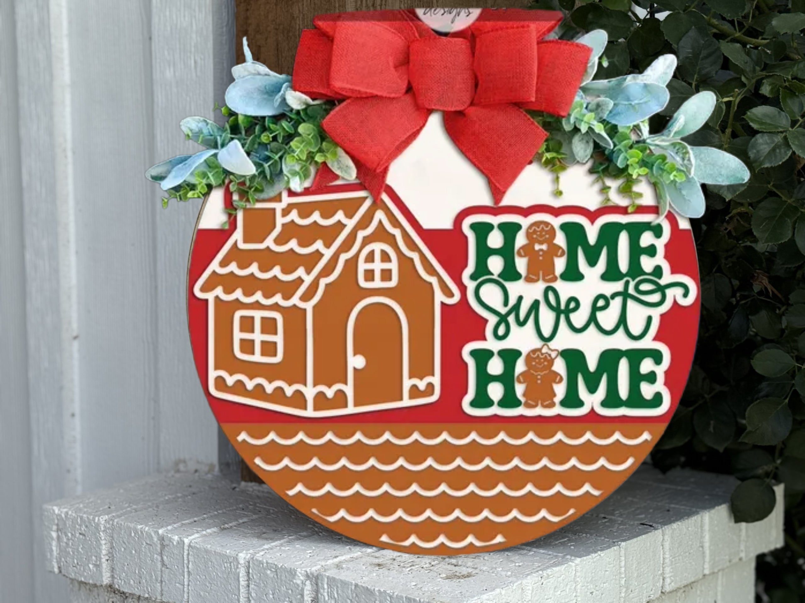 A round wooden sign decorated with a red bow, greenery, a gingerbread house, and the words Home Sweet Home with gingerbread people, leaning against a white wall near some plants.
